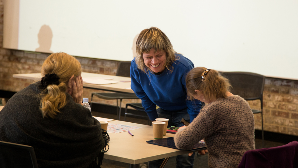 Training: Liz Martin, trainer, is wearing a blue top and is standing over a desk laughing and talking to two people