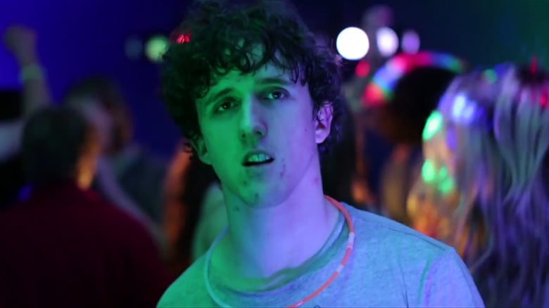 A still frame from Pulse, Daniel stands in what looks like a darkened dance floor, with a sift green light illuminating one side of his face. He wears a grey t-shirt and has a glow stick around his neck.