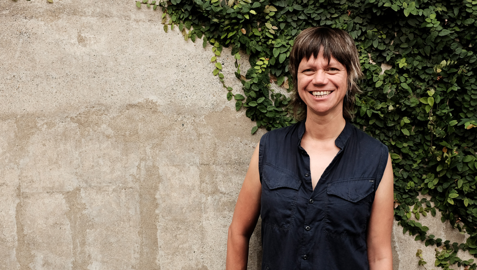 Liz Martin is standing with her back to a stone wall partially covered with a vine. Photo by Sue Wright