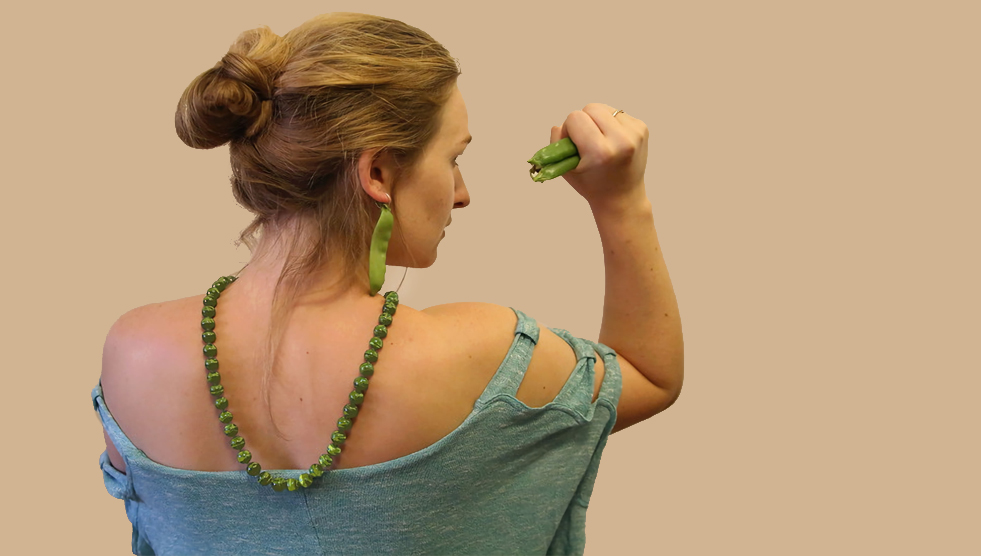 A female theatre actor wears pea earrings and a pea necklace and holds a bunch of snowpeas like a weapon