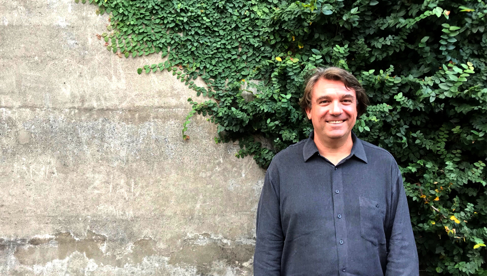 Scott O'Hara is standing with his back to a stone wall partially covered with a vine