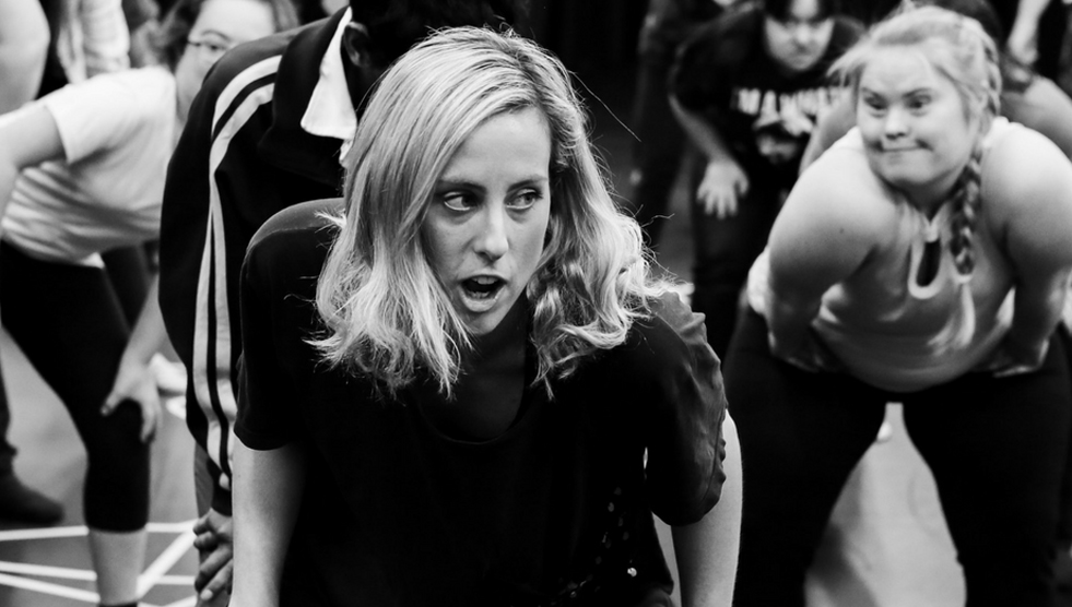 The Right Foot: A woman stands in the foreground and leads a dance workshop. Dancers are seen in the background.