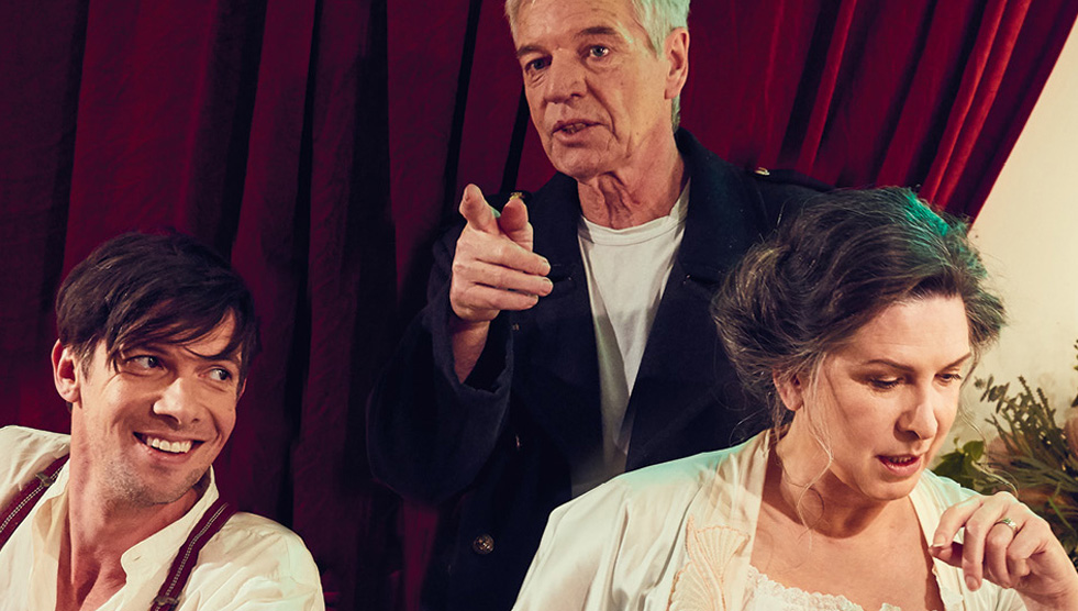 The Dance of Death: A man and a woman are sitting down. A man is standing behind them and motioning with his hand