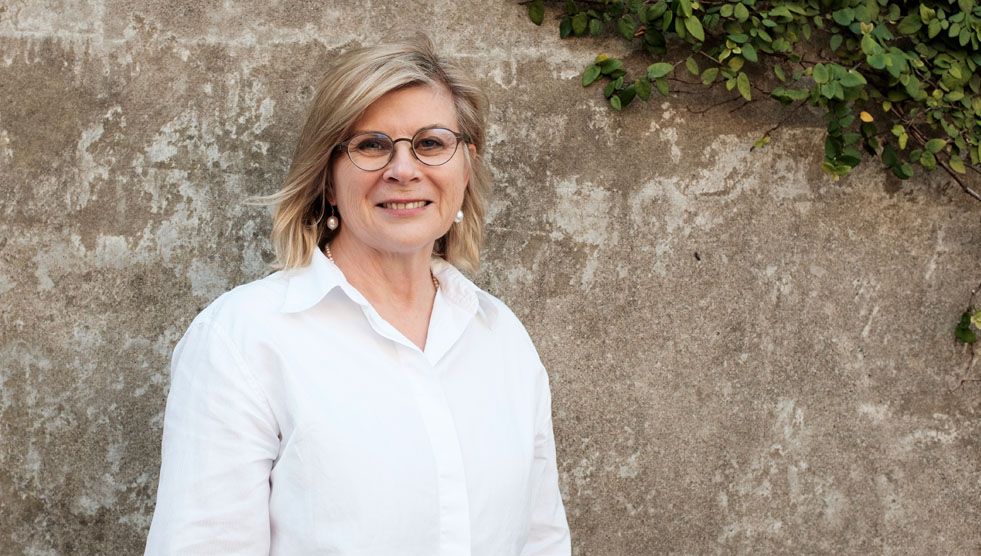 Kerry Comerford is standing with her back to a stone wall partially covered with a vine. Photo by Susan Wright