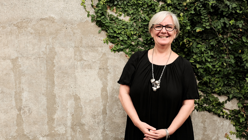 Kay Richardson is standing with her back to a stone wall partially covered with a vine. Photo by Susan Wright