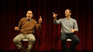 Ticket giveaway: Two men are sitting on chairs on a stage and pointing upwards with one hand