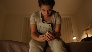A young woman is sitting on the edge of a bed and looking at an object she is holding in her hands