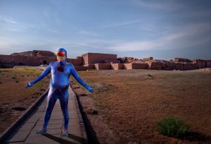 A man in a blue lycra costume with a hood is standing on a path and facing the camera