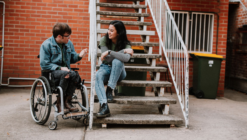 The Angus Project: A man is sitting in a wheelchair and a woman is sitting on the stairs. They are interacting.